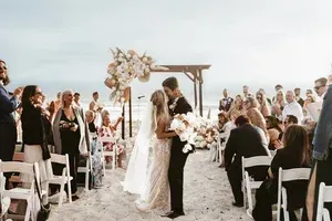 Beach wedding ceremony with bride and groom under floral arch, guests seated on white chairs at sunset