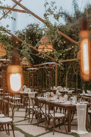 Outdoor dining table set under a wooden canopy with hanging lights and lush greenery.
