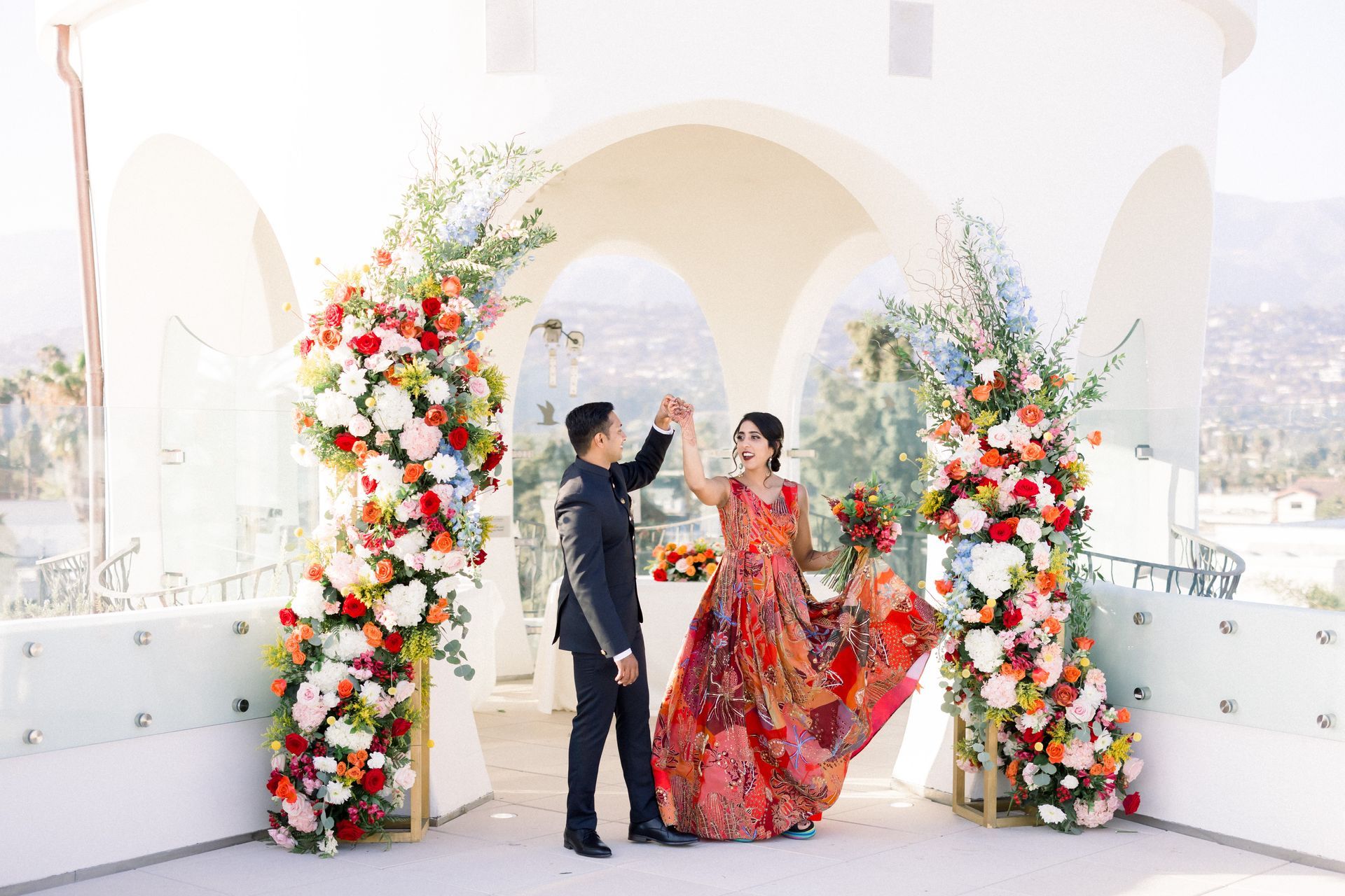 Couple dancing under floral arches at a bright rooftop wedding venue, with the woman in a red dress.