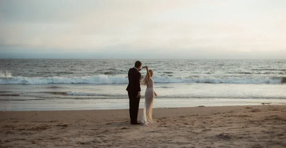 Two people standing on a sandy beach facing the ocean at sunset