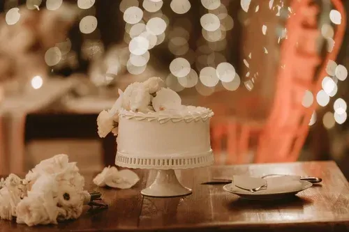 White frosted cake on a pedestal stand with flowers beside it on a wooden table, warm bokeh lights in background