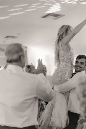 Two people lift a bride in a flowing gown at a lively celebration.