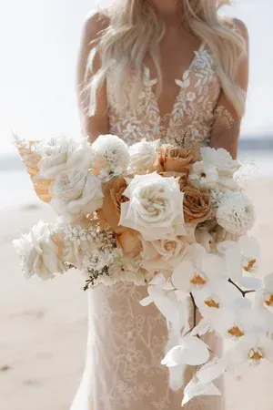 Bride holding a white and beige cascading bouquet in a lace wedding dress at the beach