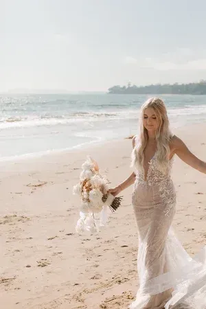 Bride in a white lace gown walking barefoot on a beach, holding a bouquet near the ocean