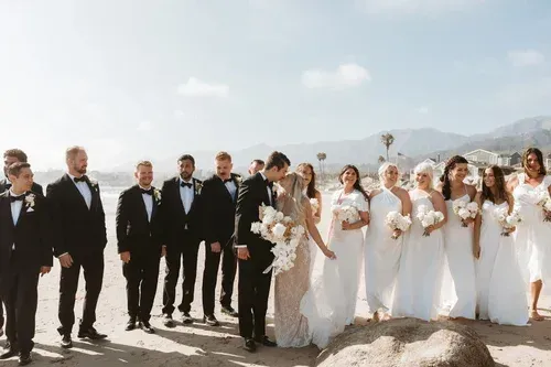 Wedding party standing on a beach with mountains in the background, bride in white and groomsmen in black tuxedos