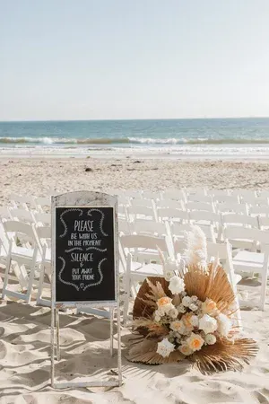 Beach wedding setup with white chairs, chalkboard sign, and dried floral arrangement by the shore