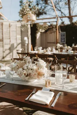 Sunlit outdoor wedding reception table with white floral centerpieces, glassware, and folded napkins