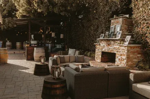 Outdoor patio lounge with wicker sofas, barrel tables, and a stone fireplace under trees