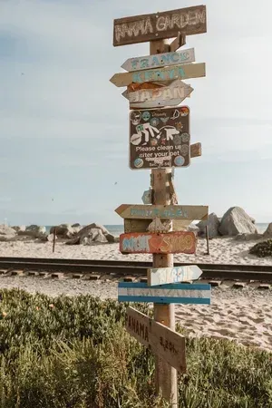 Weathered wooden signpost with colorful directional arrows beside railroad tracks and rocks on a cloudy beach.