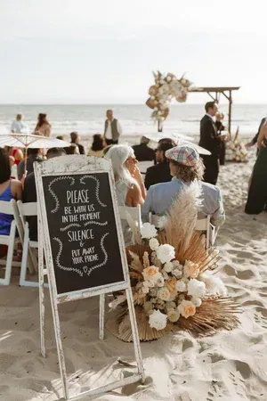 Beach wedding reception with chalkboard sign, white chairs, and floral aisle decor by the ocean