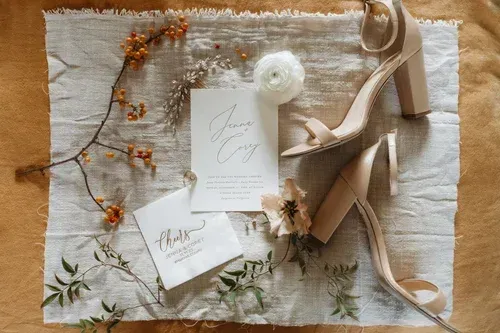 Flat lay of wedding stationery, tan heels, and white flower on a rustic neutral fabric background