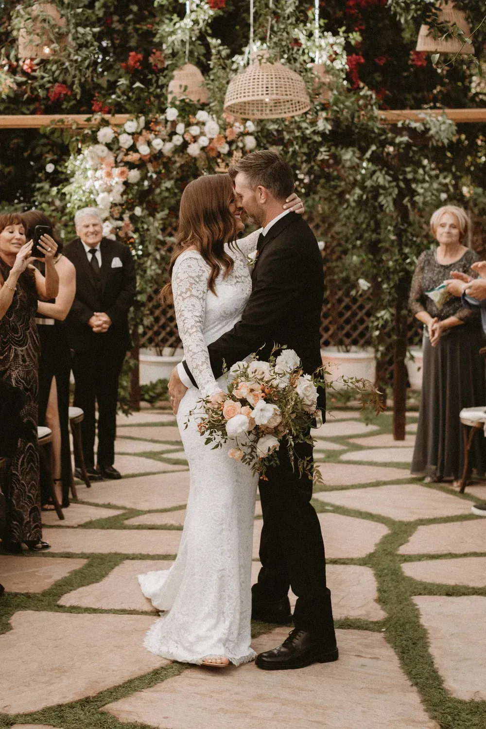 Wedding couple kissing beneath a floral arch, bride in lace dress holding bouquet, guests standing nearby