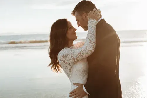 Couple embracing on a beach at sunset, facing each other in warm light.