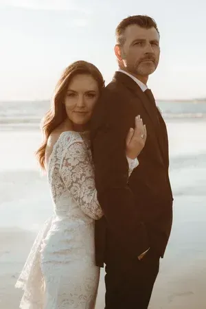 Couple standing on a beach at sunset, the woman in a white lace dress leaning against the man in a dark suit.
