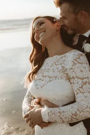 Couple embracing on a beach at sunset, with the woman in a white lace dress smiling.