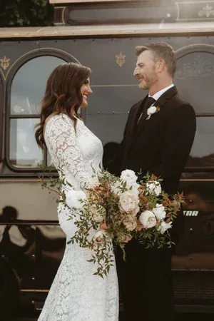 Couple in wedding attire smiling beside a dark vintage van, holding a white floral bouquet.