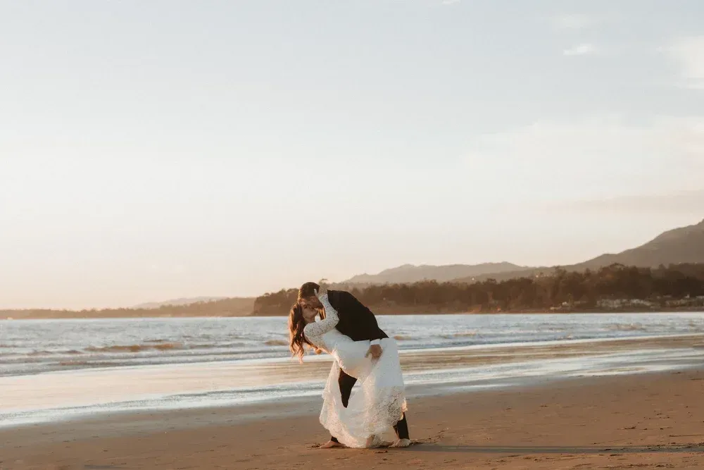 Couple embracing on a beach at sunset, with waves and a distant shoreline.