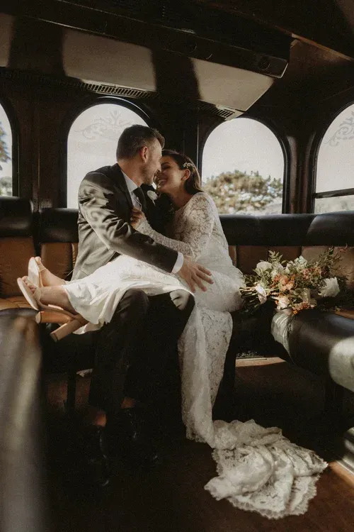 Couple embracing in a vintage car, with a bouquet and long wedding dress trailing beside them.