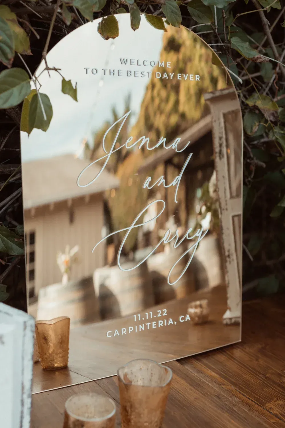 Wedding welcome sign on wooden table with candles and greenery, reading “Jennie and Larry” and “Carpinteria”