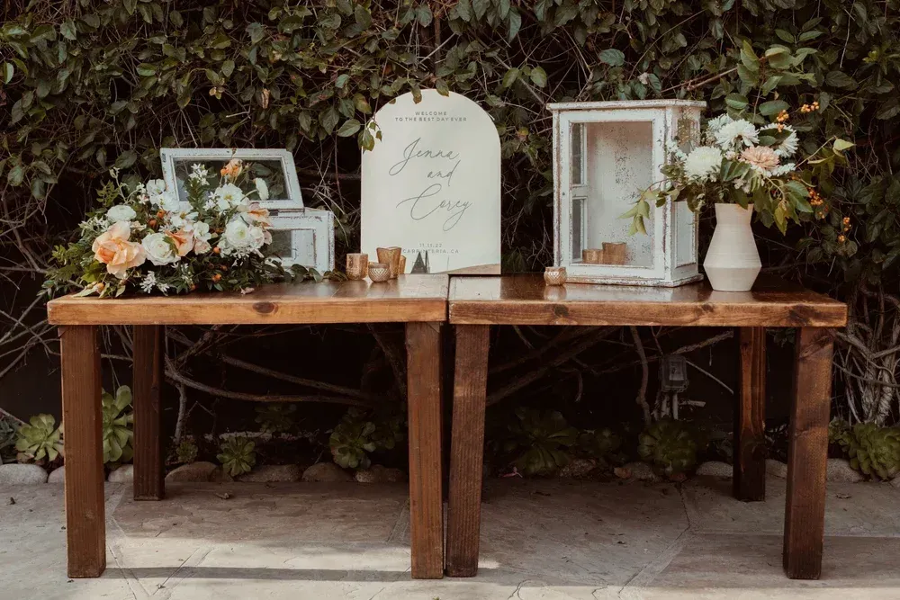 Wooden table with framed signs, flowers, candles, and decor set against a leafy hedge outdoors