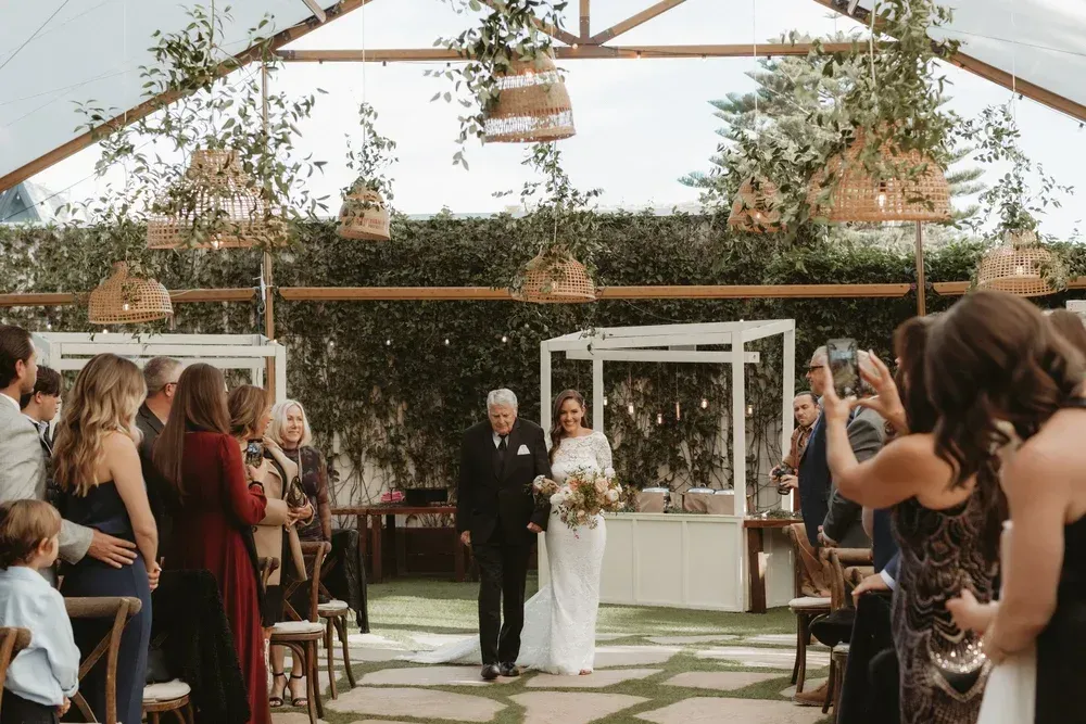 Wedding ceremony in a greenhouse with bride and groom under a floral arch, guests seated on both sides