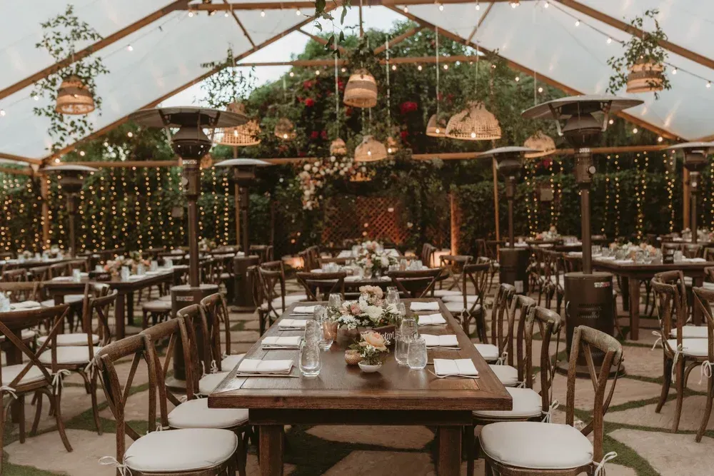 Rustic greenhouse dining setup with long wooden tables, chairs, hanging lights, and floral centerpieces