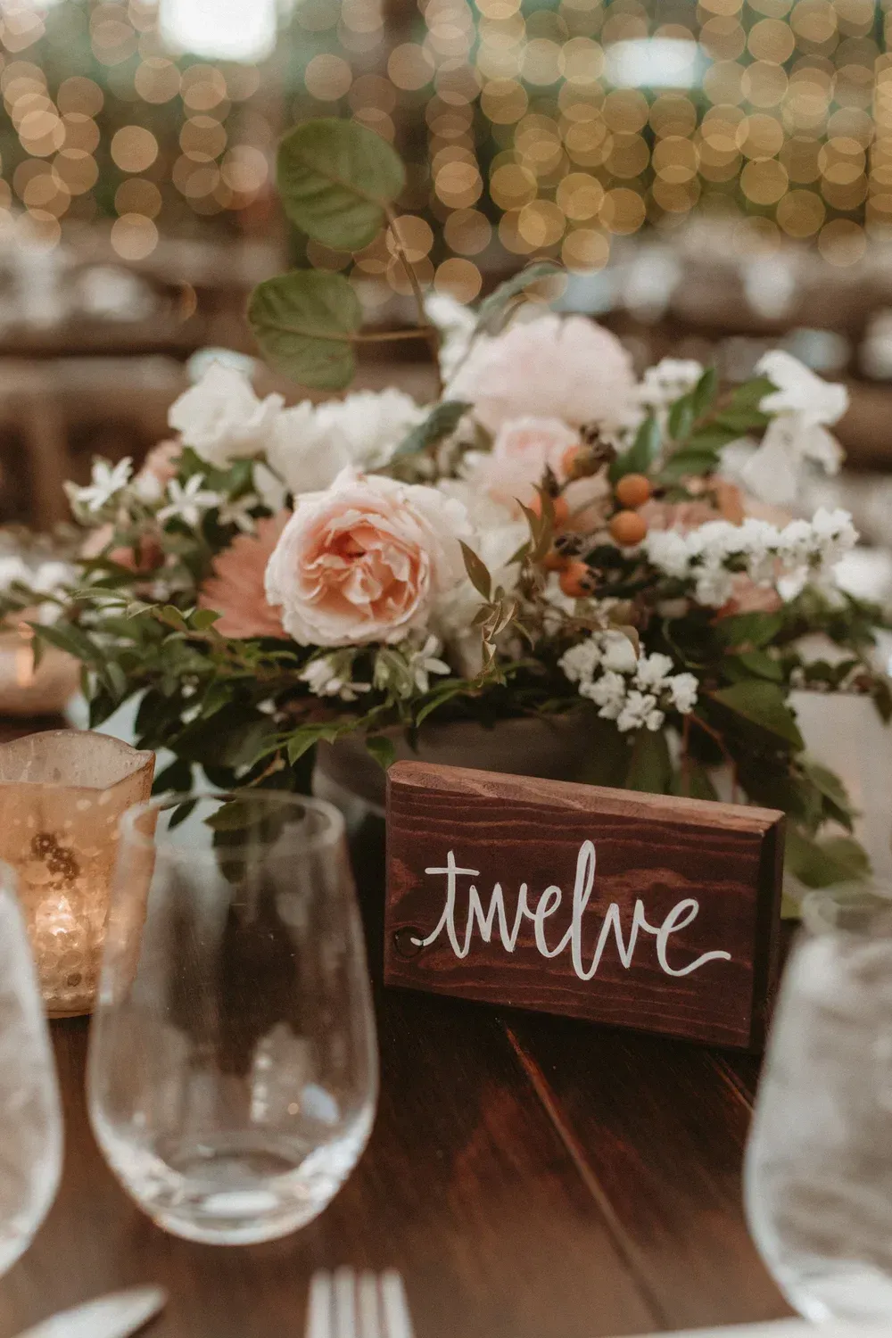 Floral table centerpiece with candles and a wooden “twelve” table sign at a wedding reception