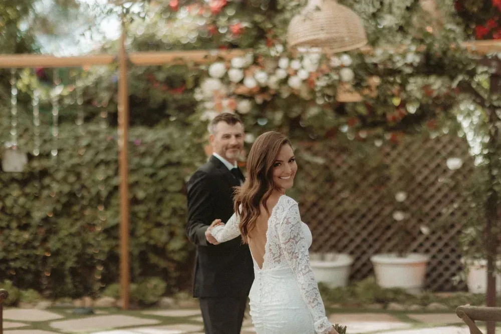 Couple dancing outdoors in a garden, the woman in a white lace dress and the man in a black suit.