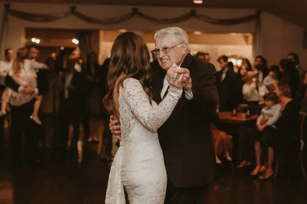 Couple dancing slowly at a warmly lit indoor celebration, smiling amid guests nearby
