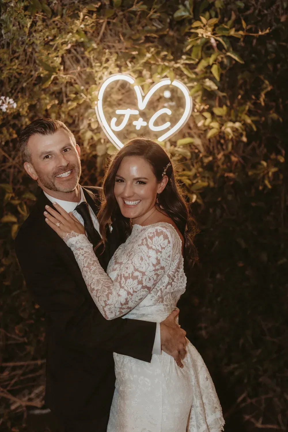 Smiling couple embracing in wedding attire under a glowing heart sign outdoors at night