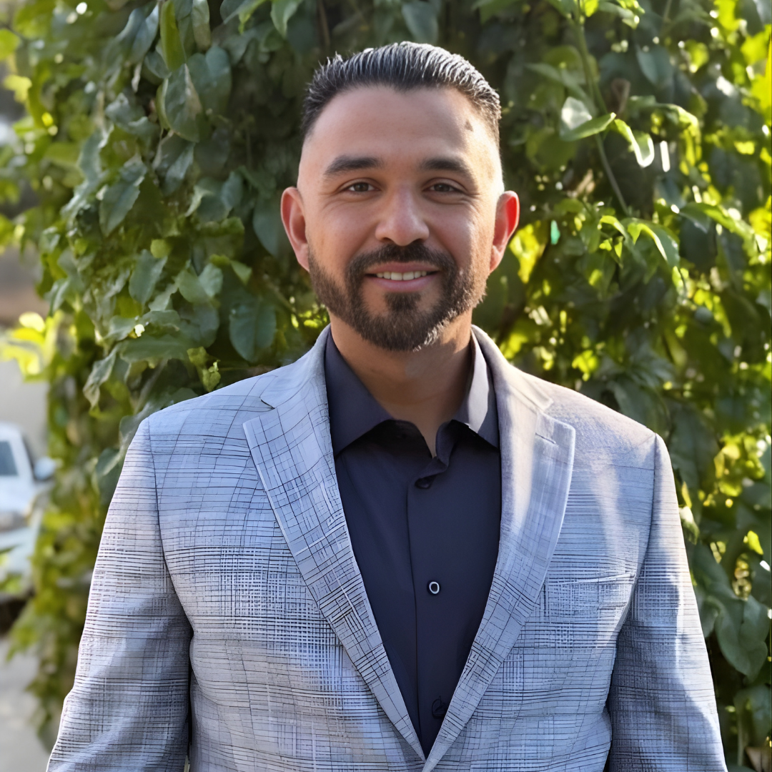 Man in a light gray blazer and navy shirt standing outdoors in front of green foliage, smiling.