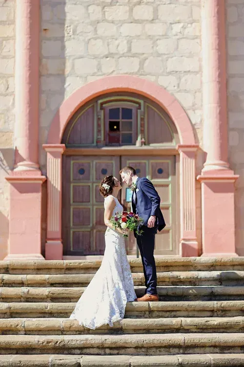 Bride and groom kissing on the steps of a pink stone building with arched doorway