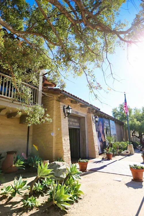 Sunlit courtyard with adobe buildings, potted plants, and a large tree casting shade