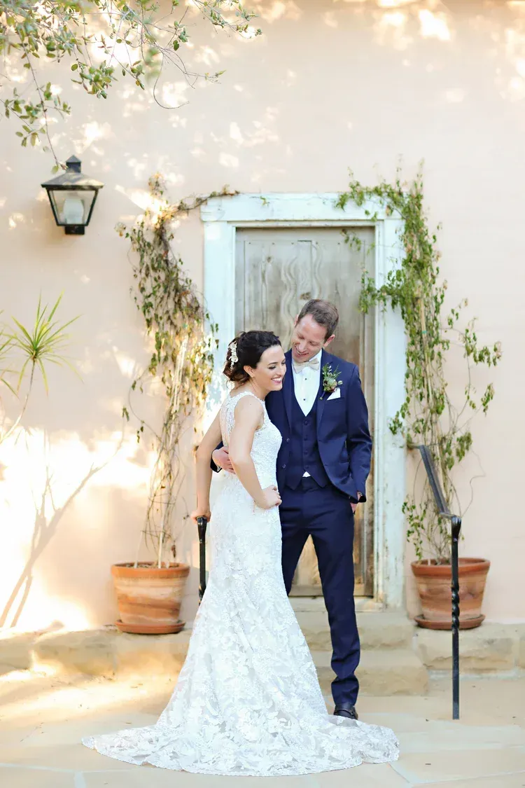 Bride and groom in wedding attire pose by a flower-adorned doorway outdoors