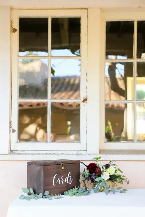 Rustic wedding welcome display on a table beneath white window panes, with flowers and a wooden sign.