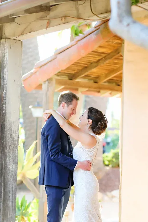 Couple embracing under a rustic wooden roof, the woman in a white dress and the man in a navy suit