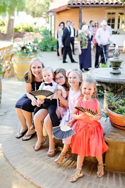 Woman and four children seated outdoors, smiling and holding paper fans near a fountain.