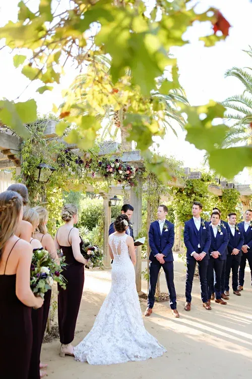 Wedding ceremony outdoors with bride in white gown facing groom and groomsmen under leafy archway