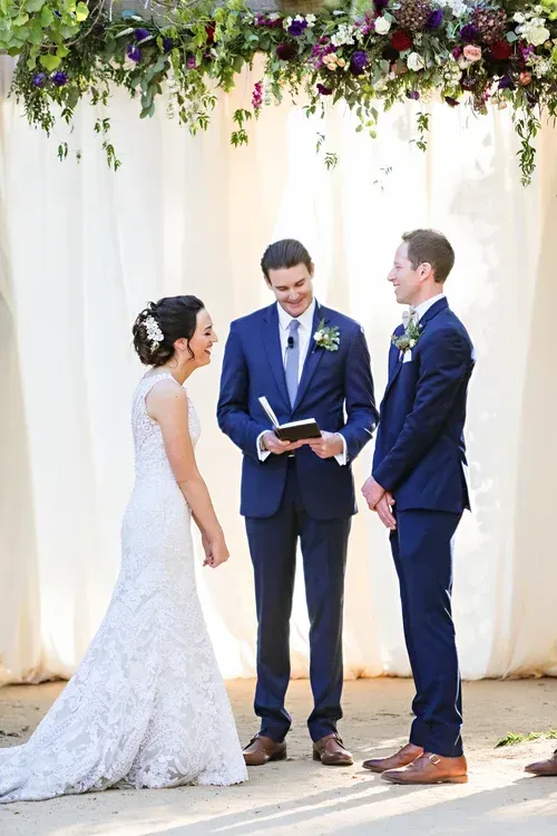 Bride and two grooms standing at a wedding ceremony under floral arch, one officiating, smiling outdoors