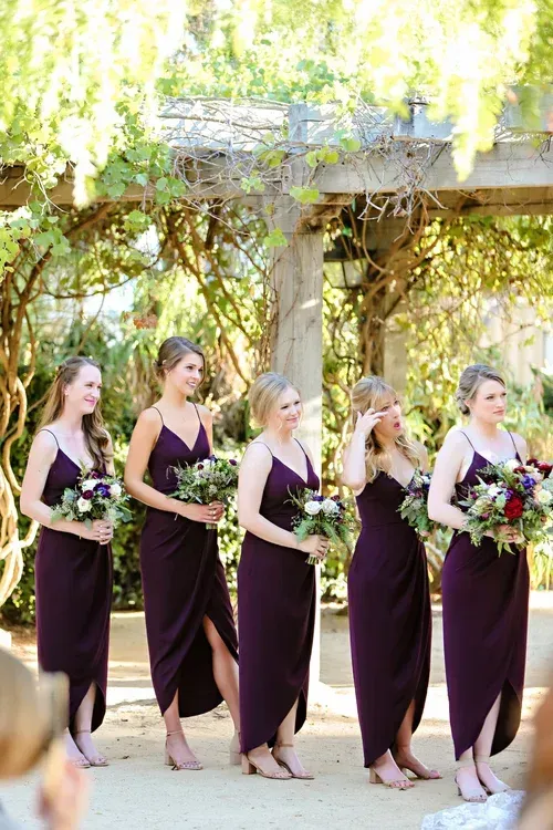 Five bridesmaids in burgundy dresses walking outdoors under a vine-covered pergola, holding bouquets