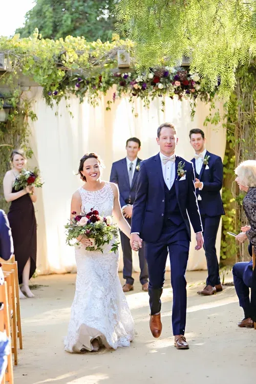 Bride and groom walking down an outdoor aisle with wedding guests under greenery