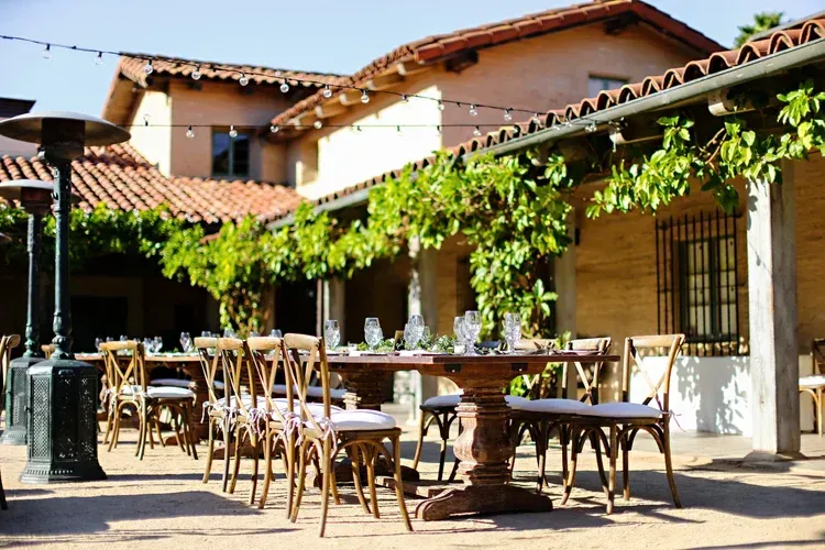 Outdoor café patio with empty tables and chairs under a vine-covered pergola beside a stucco building