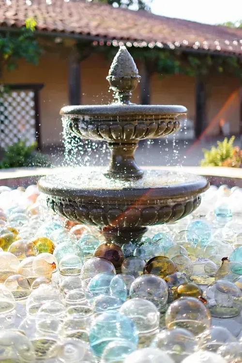 Stone fountain with water trickling into a basin surrounded by clear glass marbles in a sunlit courtyard