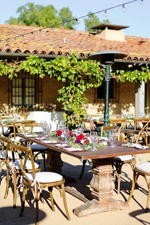 Outdoor patio dining area with a long table, wooden chairs, and vine-covered building under a tiled roof.