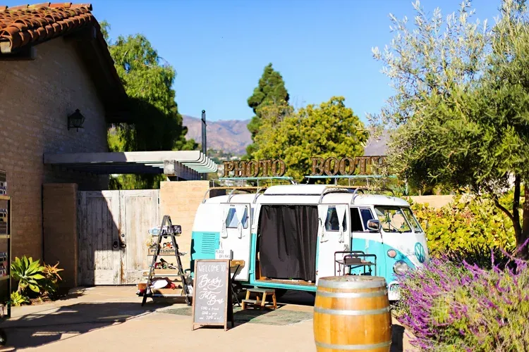 White van parked beside a house in a sunny yard with a sign, barrel, and purple flowers.