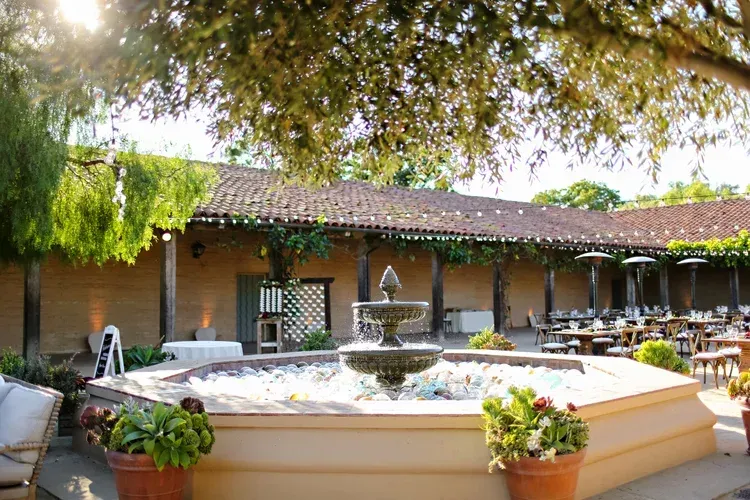 Courtyard with a tiered fountain, potted plants, and a shaded adobe building under leafy trees
