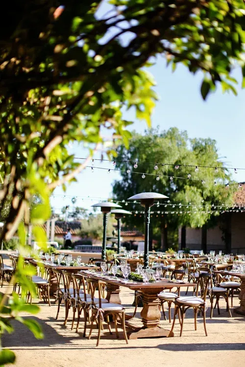 Outdoor café patio with wooden tables and chairs under trees, lit by sunlight.