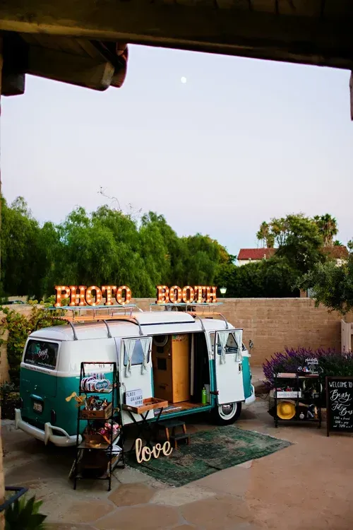 Retro photo booth van at an outdoor event, with greenery and a sign in the background.