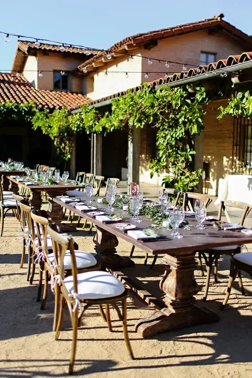 Outdoor dining tables set on a sunlit patio beside a vine-covered building with terracotta roofs.
