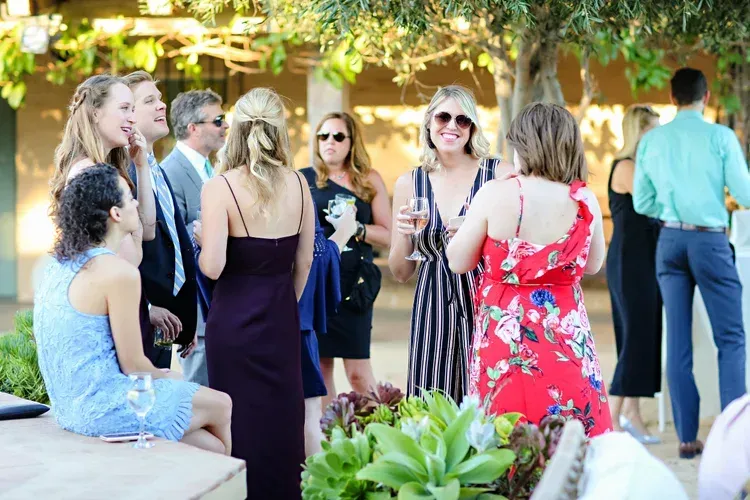 Guests mingling outdoors at a sunny social gathering, wearing colorful dresses and sunglasses